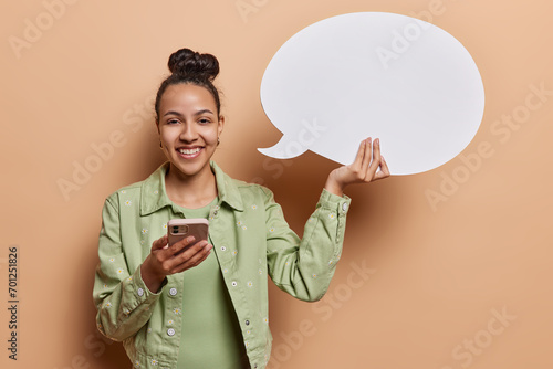 People positive emotions concept. Studio shot of young happy smiling European woman standing isolated on left on beige background wearing casual clothes holding white speech bubble with space photo