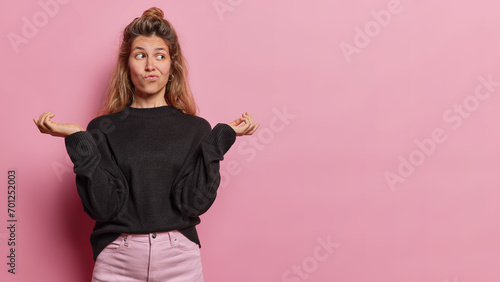 Uncertain clueless European woman spreads palms and shrugs shoulders purses lips concentrated aside dressed in casual clothes isolated over pink background copy space for your advertisement. photo