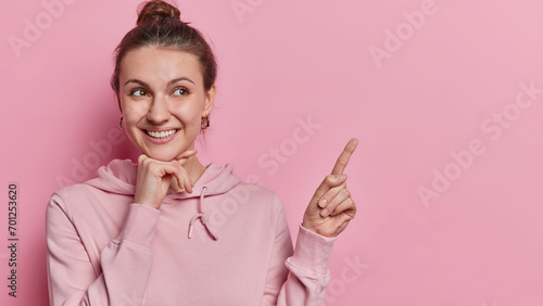 Positive good looking young woman keeps hand under chin points index finger on copy space shows banner or advertisement smiles pleasantly dressed in casual hoodie isolated over pink background photo
