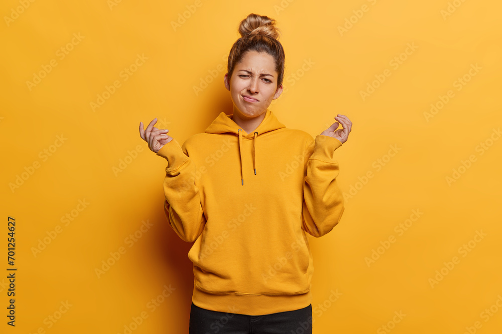 People emotions concept. Indoor photo of young confused European girl wearing yellow hoodie and black trousers standing in centre isolated spreading palms in hesitation not knowing what to do - obrazy, fototapety, plakaty 