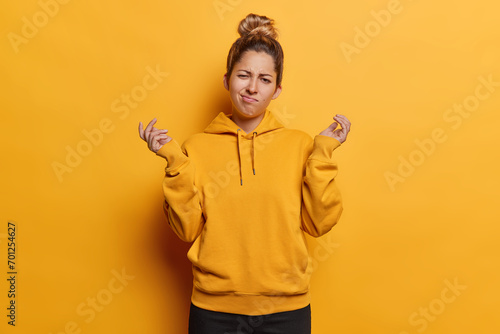 People emotions concept. Indoor photo of young confused European girl wearing yellow hoodie and black trousers standing in centre isolated spreading palms in hesitation not knowing what to do photo