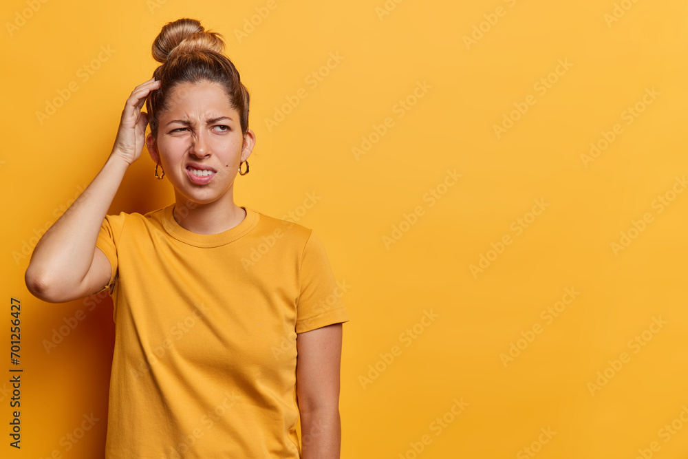 Waist up shot of European woman with hair bun scratches head and looks with bewilderment concentrated aside wears casual t shirt isolated over yellow background copy space for your promotional content Waist up shot of European woman with hair bun scratches head and looks with bewilderment concentrated aside wears casual t shirt isolated over yellow background copy space for your promotional content