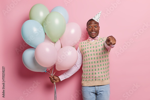 Happy surprised dark skinned bearded man points finger directly at camera holds bunch of inflated colorful balloons celebrates special occasion isolated over pink background. Hoilday concept photo