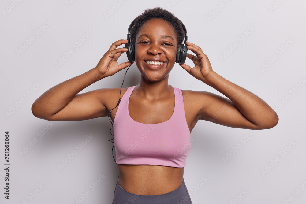 Waist up shot of slim dark skinned woman wears stereo headphones on ears enjoys favorite music has fit body smiles pleasantly isolated over white wall. Sporty female model listens song from playlist Waist up shot of slim dark skinned woman wears stereo headphones on ears enjoys favorite music has fit body smiles pleasantly isolated over white wall. Sporty female model listens song from playlist