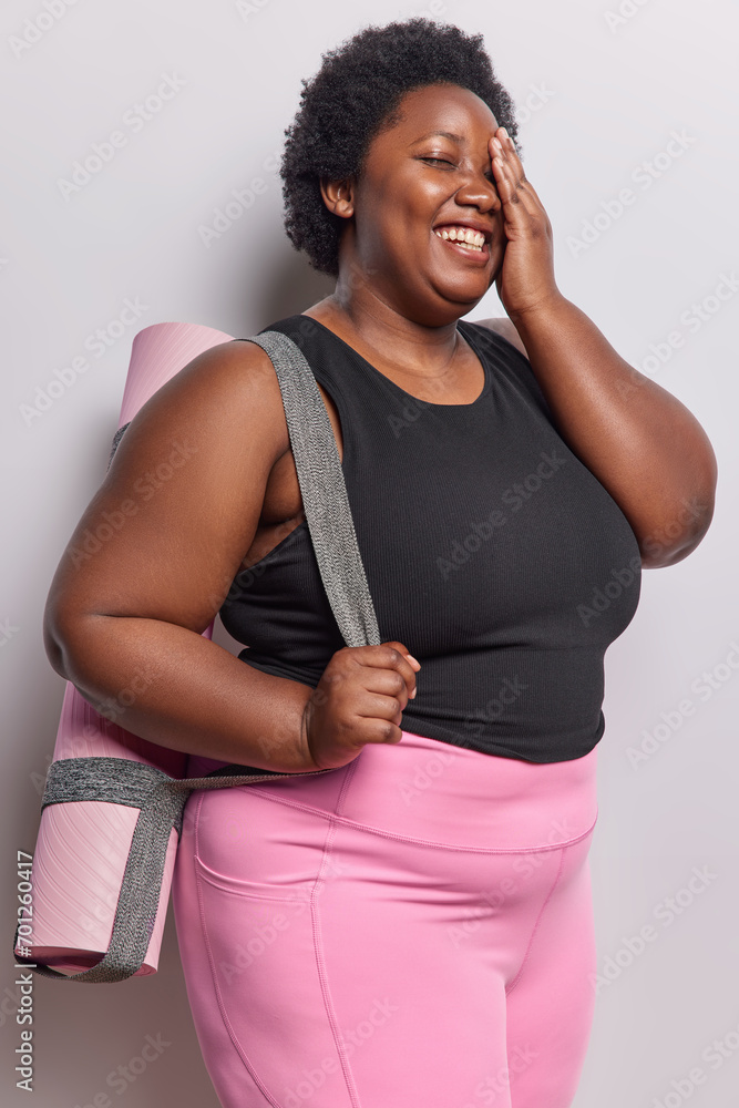 Vertical shot of plump dark skinned woman makes face palm smiles positively as gains results in loosing weight dressed in sportswear carries fitness mat going to have pilates or cardio training.