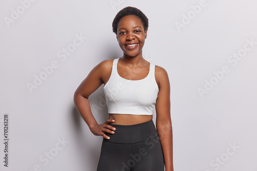 Beautiful cheerful fitness woman with short curly hair keeps hand on waist dressed in sportswear smiles gladfully has motivation to go in for sport isolated on white background. Athletic female model