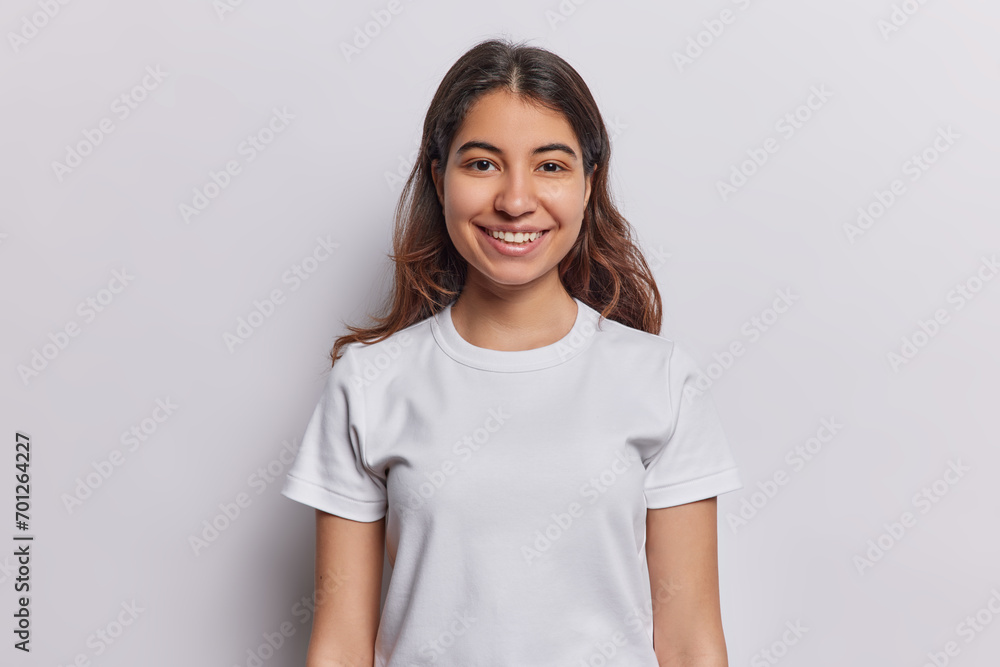 Happiness concept. Indoor waist up of young cheerful smiling Iranian girl with long brunette hair standing in centre isolated on white background keeping hands down looking straight at camera Happiness concept. Indoor waist up of young cheerful smiling Iranian girl with long brunette hair standing in centre isolated on white background keeping hands down looking straight at camera