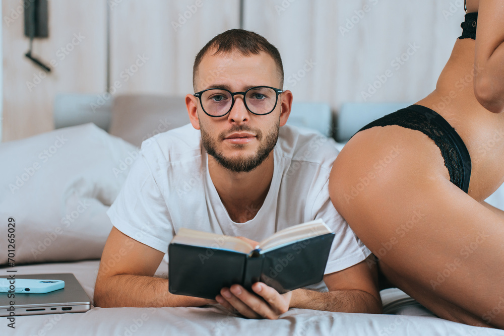 Man in glasses reading a book in bed, with a woman in lingerie beside him, suggesting a relaxed yet intimate atmosphere. Man in glasses reading a book in bed, with a woman in lingerie beside him, suggesting a relaxed yet intimate atmosphere.