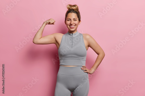 Positive European woman flexing biceps and smiles happily dressed in sports clothing smiles gladfully maintaines active lifestyle for good health poses over pink background feels proud of achievements
