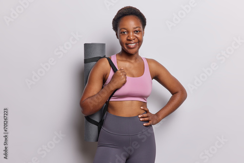 Horizontal shot of active dark skinned woman being in good physical shape wears tracksuit keeps hand on waist smiles happily leads active lifestyle poses with rolled fitness mat on shoulder indoor