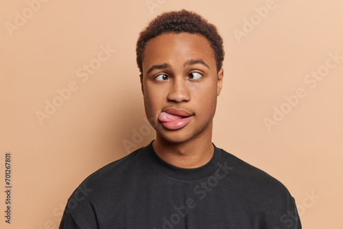 Studio close up of young funny African american man with short curly hair wearing black t shirt standing in centre isolated on beige background keeping hands down showing tongue making grimace photo
