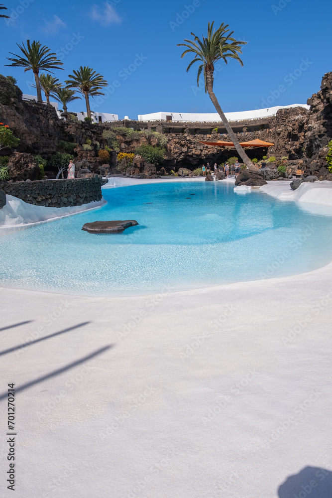 Pool of blue waters and palemeeras, outside the cave of Los Jameos del Agua. Light at the end of the cave. Sky with big white clouds. Lanzarote, Canary Islands, Spain.