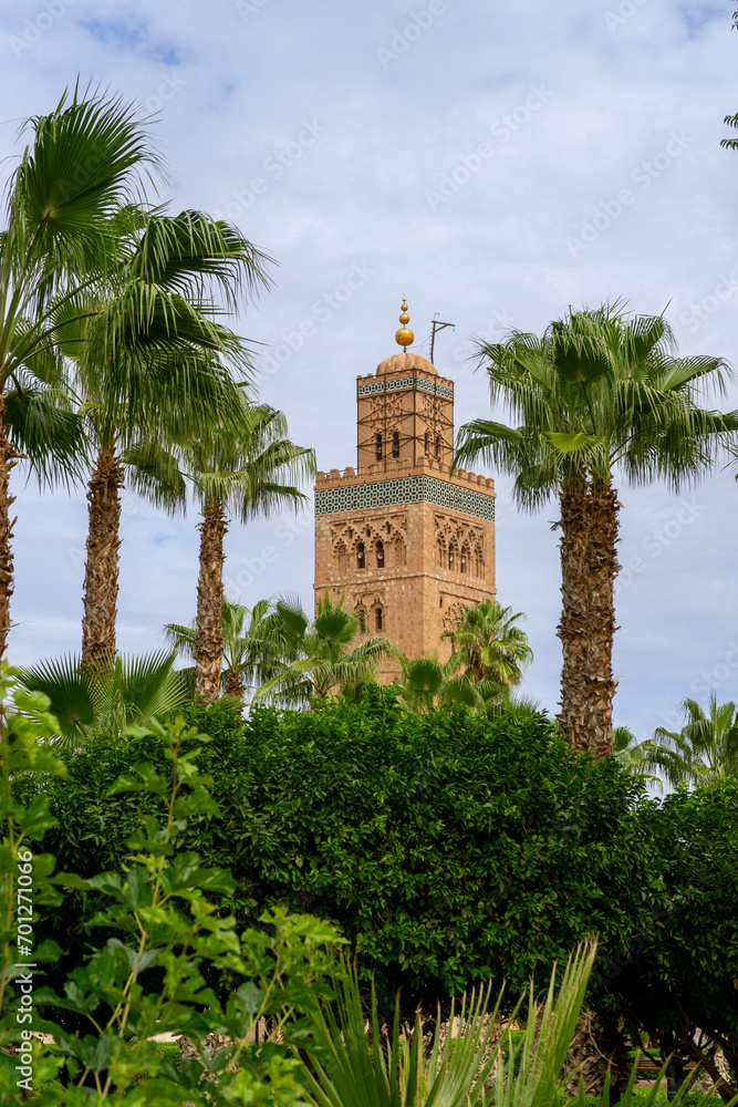 Photo of the upper part of the tower of the Koutoubia mosque in the city of Marrakech, tower covered by trees and palm trees. Cloudy day.