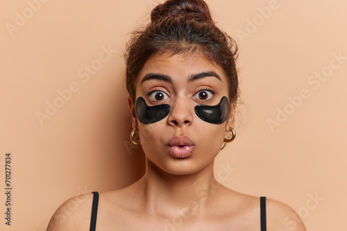 Beauty procedure. Studio close up of young beautiful European lady standing in centre isolated on beige background applying black patches to reduce wrinkles keeping long hair in bun wearing undershirt