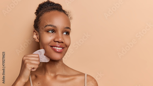 Cosmetic natural trend. Pretty dark skinned woman with dark hair gathered in bun smiles tenderly uses gua sha quartz stone for massage focused aside poses against brownn background copy space photo