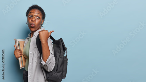 Photo of impressed curly haired student carries notepads carries black rucksack points thumb on copy space for your promotional content enjoys studying at college isolated over blue background photo