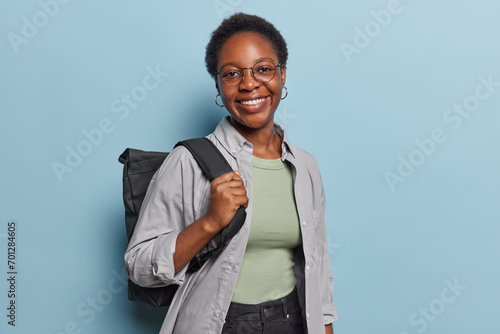 Indoor shot of pretty dark skinned schoolgirl smiles happily returns home after studying carries black rucksack wears shirt and trousers isolated over blue background. Studying and education concept photo