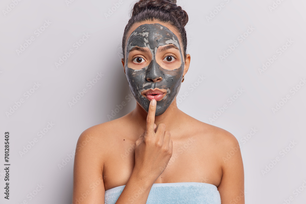 Photo of impressed Latin woman with dark hair keeps finger on lips applies facial clay mask for skin treatment wrapped in bath towel isolated over white background. Beauty procedures at home Photo of impressed Latin woman with dark hair keeps finger on lips applies facial clay mask for skin treatment wrapped in bath towel isolated over white background. Beauty procedures at home