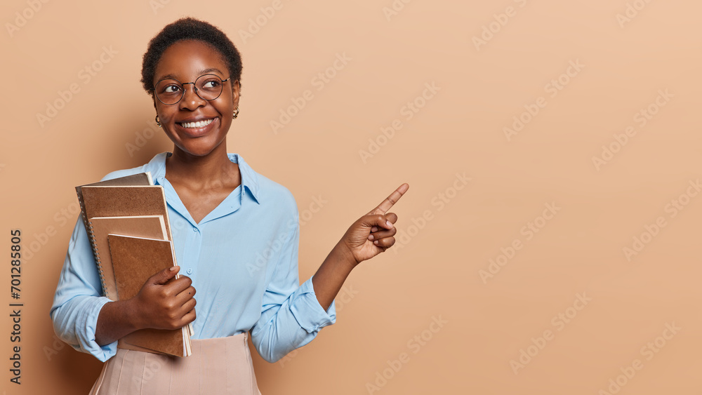 Pleased smiling dark skinned female student wears round spectacles formal blue shirt and skirt holds spiral notebooks and textbooks points index finger on copy space for your promotional content Pleased smiling dark skinned female student wears round spectacles formal blue shirt and skirt holds spiral notebooks and textbooks points index finger on copy space for your promotional content