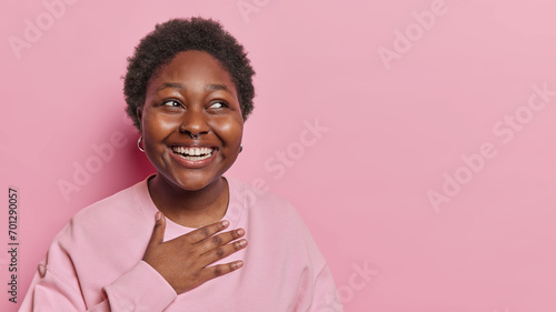 Happy positive dark skinned woman smiles gladfully keeps hand on chest giggles as notices something funny dressed in casual sweatshirt has piercing in nose isolated over pink background copy space