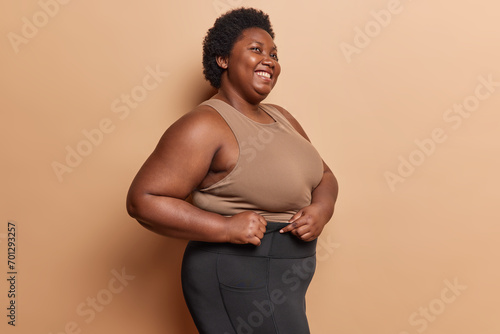 Sideways shot of cheerful overweight woman dressed in t shirt and leggings smiles broadly going to have fitness for weight loss stands indoors against brown background. Sport and motivation concept