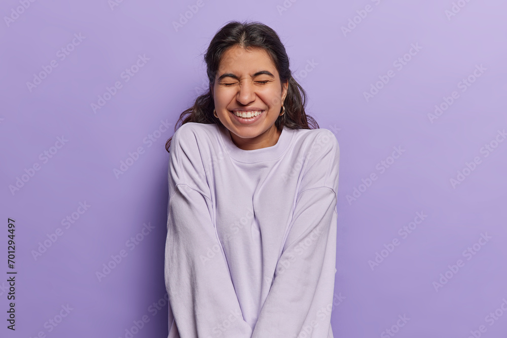 Waist up shot of positive Iranian woman with dark hair smiles happily keeps eyes closed recalls something funny dressed casually isolated over purple background. Positive human emotions concept Waist up shot of positive Iranian woman with dark hair smiles happily keeps eyes closed recalls something funny dressed casually isolated over purple background. Positive human emotions concept