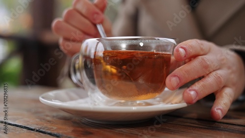 Woman adds sugar to black tea into clear cup at table in cafe. Relaxed woman sweetening tea in cafe in cold weather. Cozy ambiance and sipping hot tea with sugar prepared by woman in cafe of park