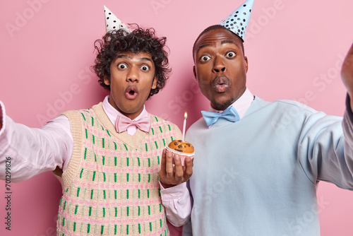Photo of shocked stupefied young men stand closely to each other and keeps arms outstetched hold delicious cupcake with burning candle isolated over pink background. Birthday celebration concept