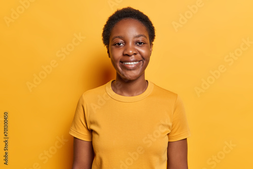 Horizontal shot of good looking millennial girl with curly hair happ smile on face expresses positive emotions dressed in casual t shirt looks gladfully at camera isolated over yellow background. © Wayhome Studio