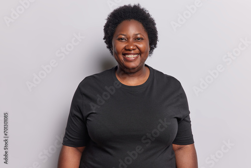 Portrait of cheerful plump African woman with curly hair smiles gladfully dressed in casual black t shirt looks directly at camera poses against white background. People ethnicity positive emotions