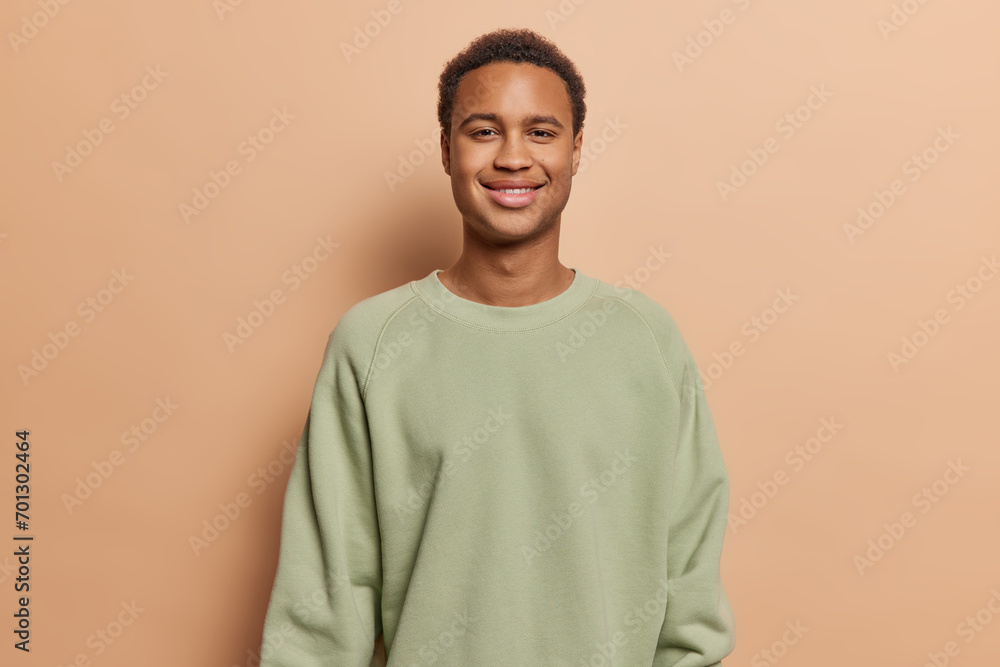 Portrait of handsome dark skinned man with short hair smiles pleasantly looks directly at camera dressed in casual sweatshirt poses against brown studio background. People and emotions concept