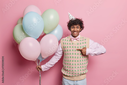 Horizontal shot of positive curly haired Hindu man points at himself smiles pleasantly asks who me wears festive clothing holds bunch of inflated colorful balloons isolated over pink background
