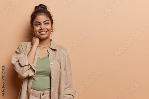 Horizontal shot of pleased tender Iranian girl keeps hand on neck smiles happily concentrated aside being in good mood has pleasant thoughts in mind dressed in stylish clothes isolated over brown wall