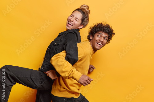 Positive female and male friends foolishing around gve piggyback ride to each other laugh happily dressed in black and yellow clothes pose indoor. Girlfriend and boyfriend spend free time together photo