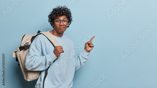 People emotions concept. Indoor sideway waist up of young dissatisfied Hindu male student standing on left isolated on blue background wearing casual clothes pointing at blank space for your promotion photo