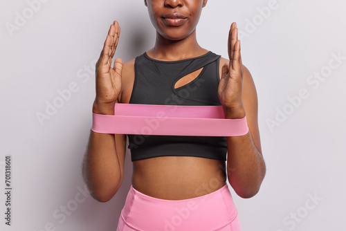 Cropped shot of dark skinned unrecognizable woman exersises with elastic resistance band has workout at home uses sport equipment dressed in black cropped top and leggings isolatedd over white wall photo