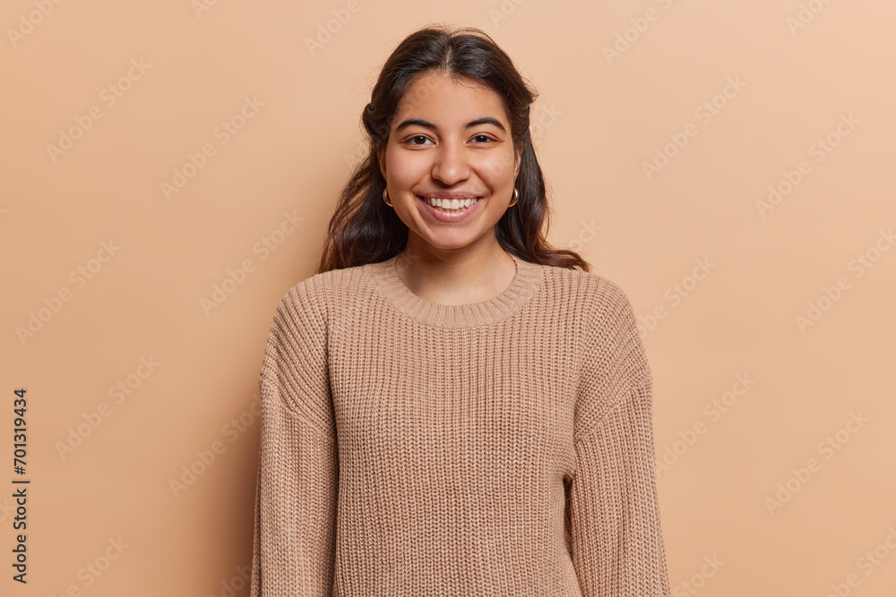 Portrait of pleasant looking Iranian girl with toothy smile has good mood dressed in casual knitted jumper looks directly at camera isolated over brown background. People and positive emotions concept Portrait of pleasant looking Iranian girl with toothy smile has good mood dressed in casual knitted jumper looks directly at camera isolated over brown background. People and positive emotions concept