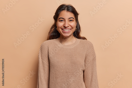 Portrait of pleasant looking Iranian girl with toothy smile has good mood dressed in casual knitted jumper looks directly at camera isolated over brown background. People and positive emotions concept photo