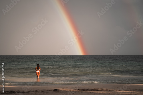 A girl in a bathing suit stands on the beach in front of a rainbow.