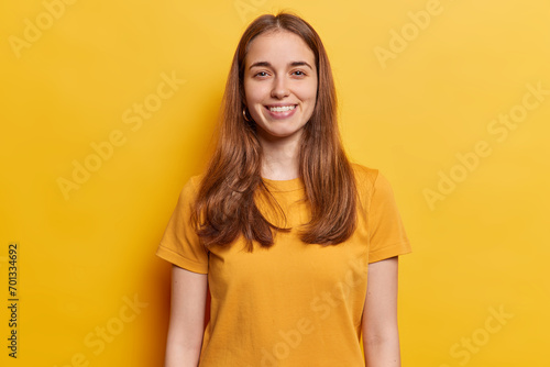 Waist up shot of pretty young woman with long straight hair smiles gladfully dressed in casual t shirt stands happy indoor isolated over yellow background. People and positive emotions concept