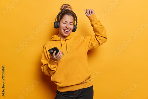 Carefree joyful young woman enjoying awesome beat in new stereo headphones shakes arms and exclaims loudly closes eyes from pleasure dressed in casual hoodie isolated over vivid yellow background.