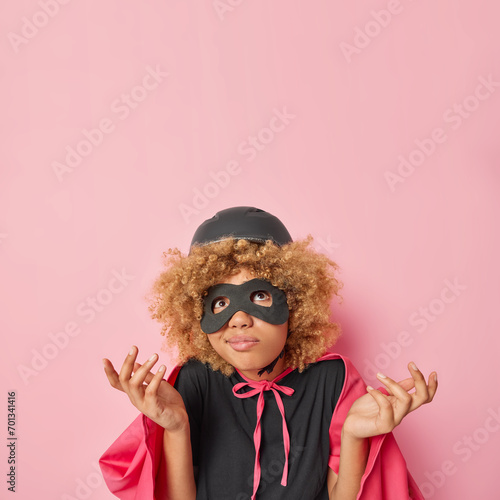 Vertical shot of hesitant female superhero spreads palms and concentrated above with clueless expression wears helmet helmet eyemask and cloak isolated over pink background copy space for your promo photo