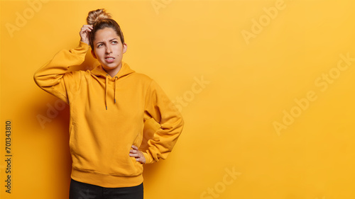 Photo of confused hesitant European woman scratches head looks embarrassed aside tries to solve problem wears casual hoodie isolated over yellow background copy space for your advertising content