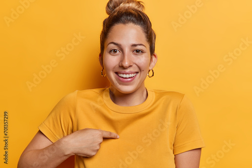 Positive European woman with hair gathered in bun points at herself asks who me being glad to be picked smiles toothily dressed in casual t shirt isolated over yellow background. Self promoting