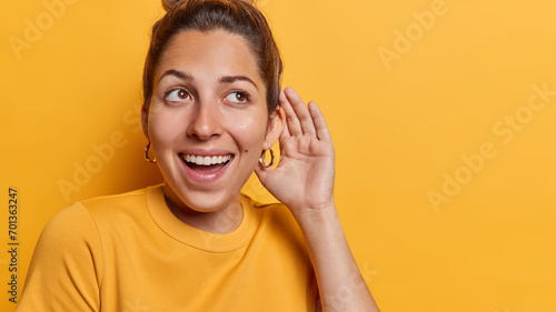 Horizontal shot of healthy young cheerful woman keeps hand near ear tries to overhear rumors listens private information with interest has curious expression isolated over yellow background. photo
