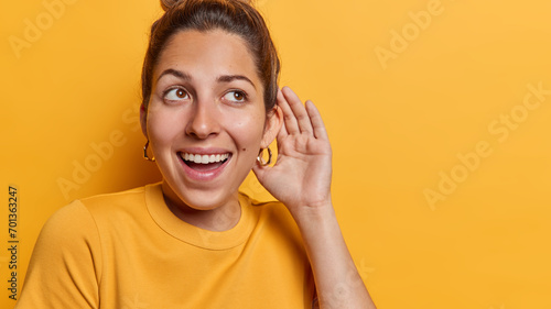 Schilderij op canvas Horizontal shot of healthy young cheerful woman keeps hand near ear tries to overhear rumors listens private information with interest has curious expression isolated over yellow background