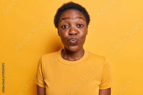 Human facial expressions. Lovely dark skinned woman keeps lips folded focused at camera has romantic mood dressed in casual t shirt isolated over vivid yellow background wants to kiss someone