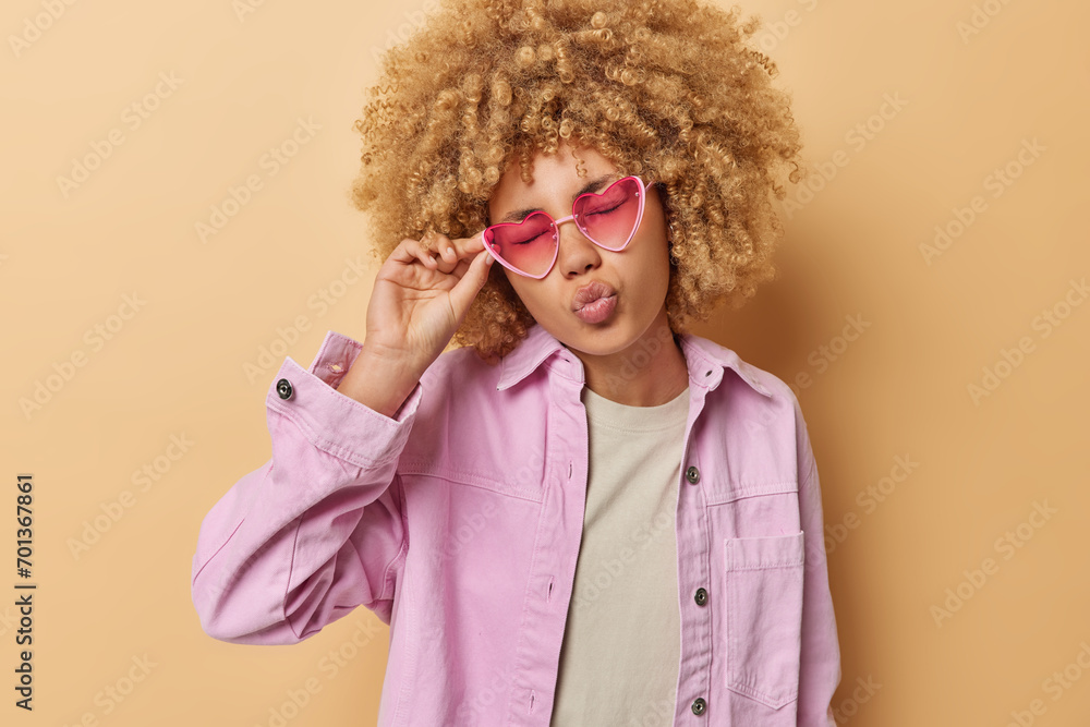 Horizontal shot of pretty curly haired woman keeps lips rounded has eyes closed romantic expression touches rim of sunglasses waits for kiss wears pink jacket isolated over brown background. Horizontal shot of pretty curly haired woman keeps lips rounded has eyes closed romantic expression touches rim of sunglasses waits for kiss wears pink jacket isolated over brown background.