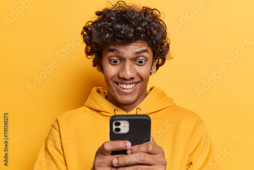 Studio photo of young glad smiling Hindu male student standing isolated in centre on yellow background holding smartphone reading message with good news or pleasant funny information on internet