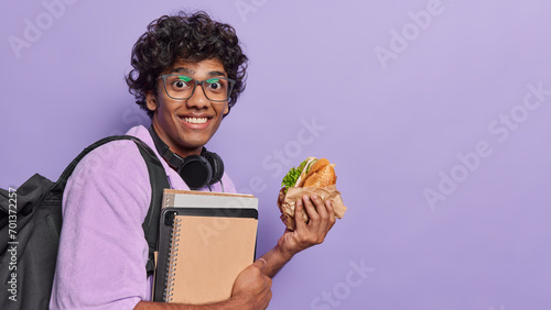 People positive emotions concept and student lifestyle. Studio sideway photo of young happy smiling Hindu male holding books and burger standing on left isolated on purple background with space